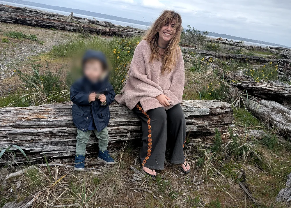 Miranda and her toddler sitting together on a driftwood log among beach grass.