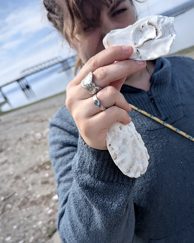 Miranda holding an oyster shell up in front of her face on the beach.